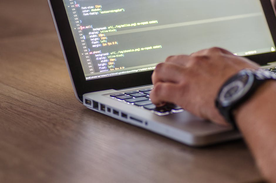 A close-up image of a person's hand typing on a silver laptop keyboard placed on a wooden desk. The screen displays lines of code or programming text in a dark background with colorful syntax highligh