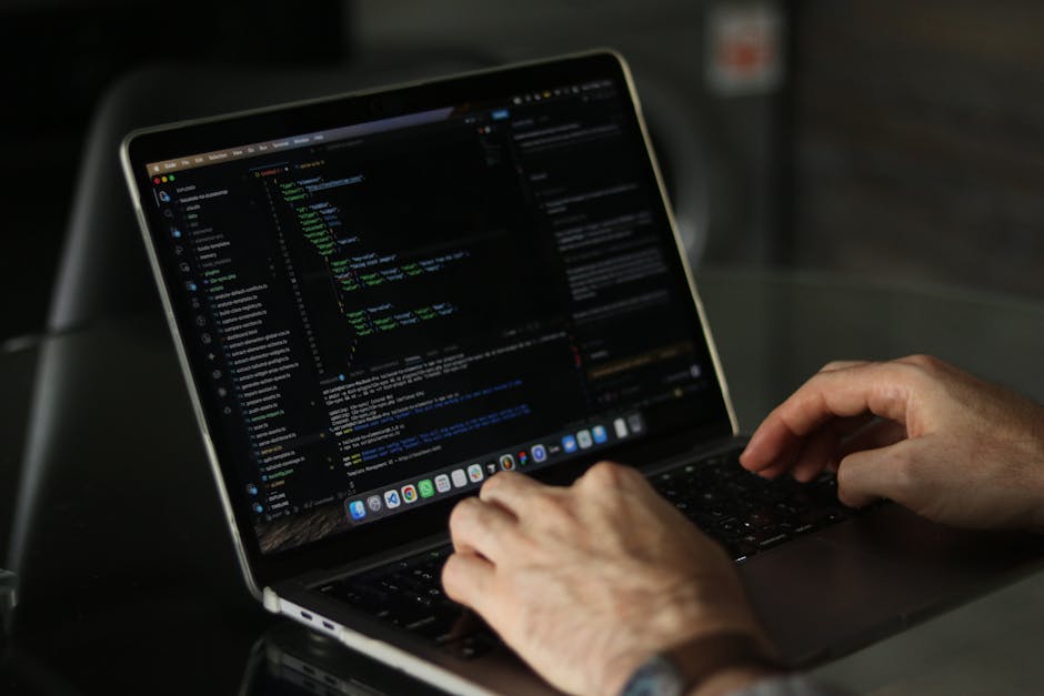 An indoor workspace featuring a computer setup with a monitor displaying lines of code, surrounded by numerous tangled cables, a keyboard, and a mouse on a dark desk. Behind the computer, there are tw